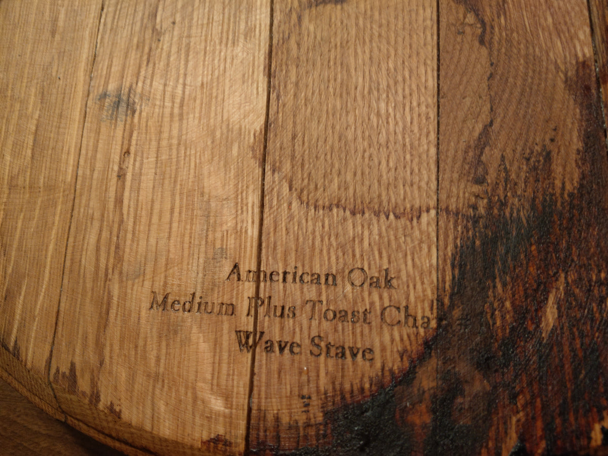 Close-up of a wooden whiskey barrel lid made from American Oak wood, with engraved text reading 'American Oak Medium Plus Toast Charcoal Wave Stove'.
