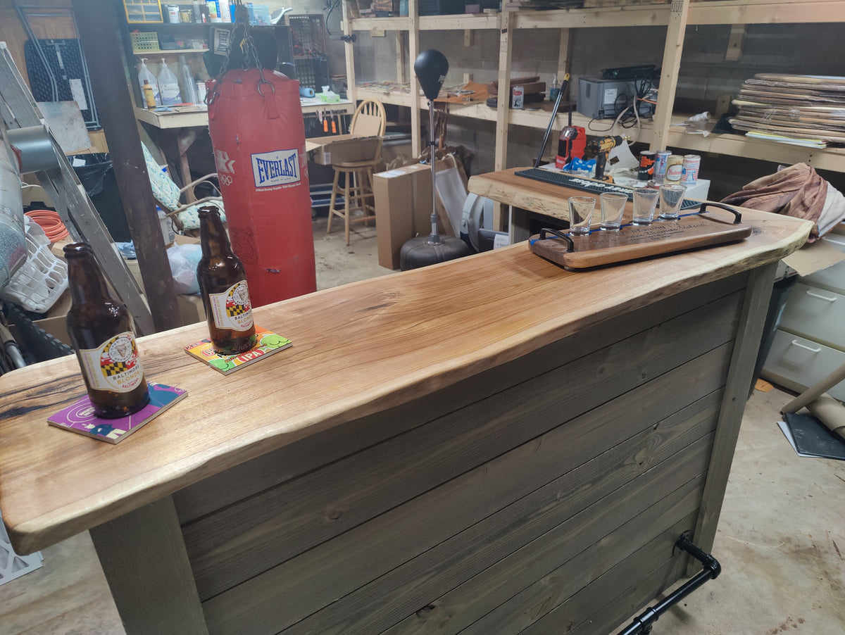 A rustic wooden bar counter with a live edge, made from stained wood panels, shown in a cluttered workshop or garage setting. Two brown beer bottles and colorful coasters sit on the left side of the counter, and a shot glass flight tray rests on the right.