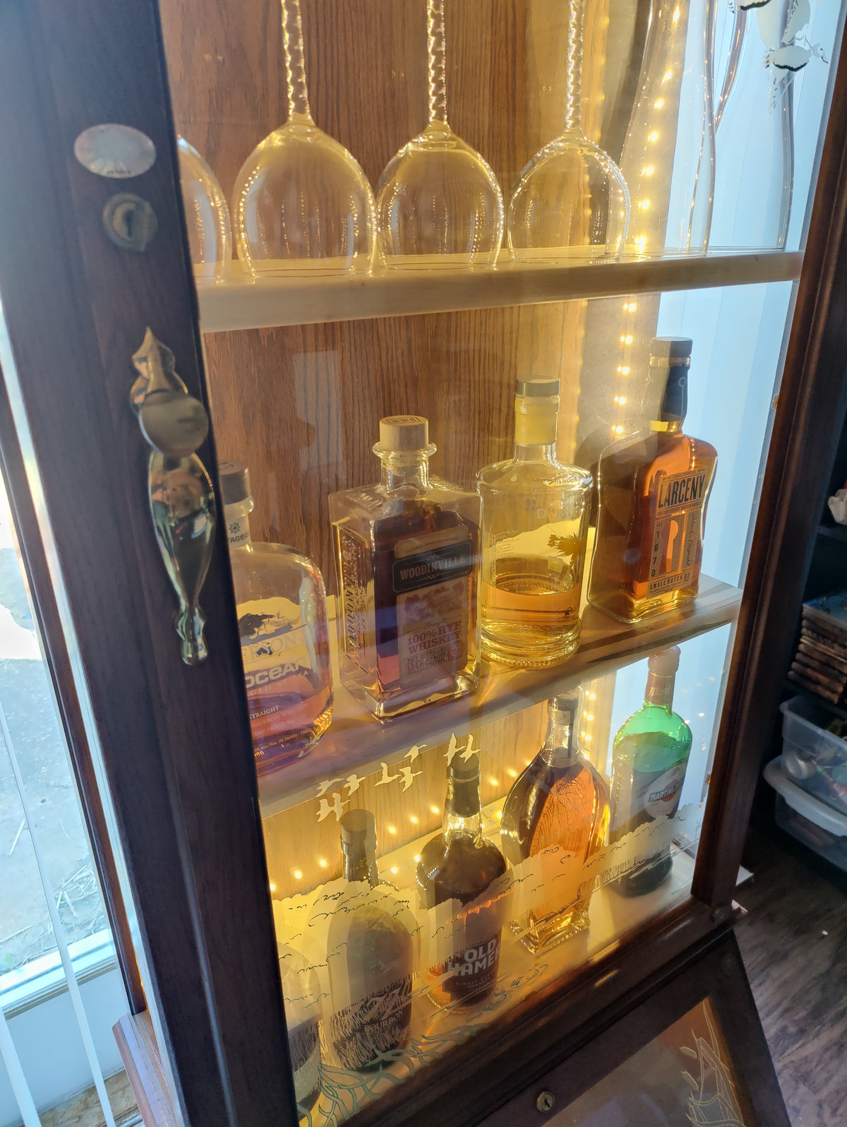 A tall wooden bar cabinet with glass doors and shelves, illuminated by warm string lights. Several liquor bottles are displayed on the shelves, and two clear wine glasses hang from chains on the top shelf.