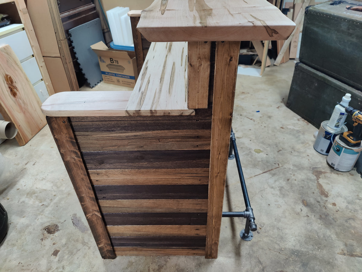 A rustic wooden bar counter or stool with a thick top, a slatted backrest made of dark and medium brown wood planks, and a black metal pipe support frame, photographed in a workshop.