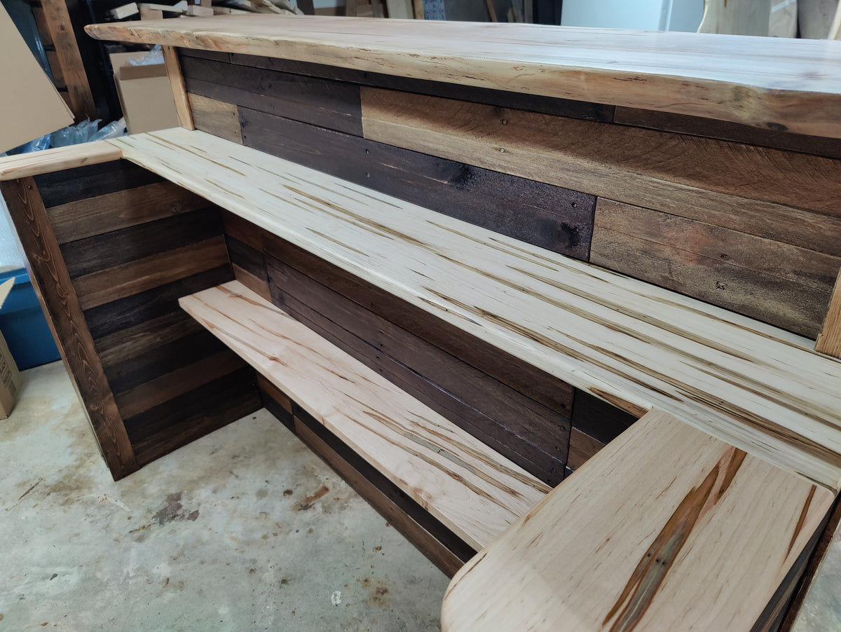 A rustic wooden bar counter with integrated shelves, featuring a mix of light and dark reclaimed wood panels and a light wood countertop.