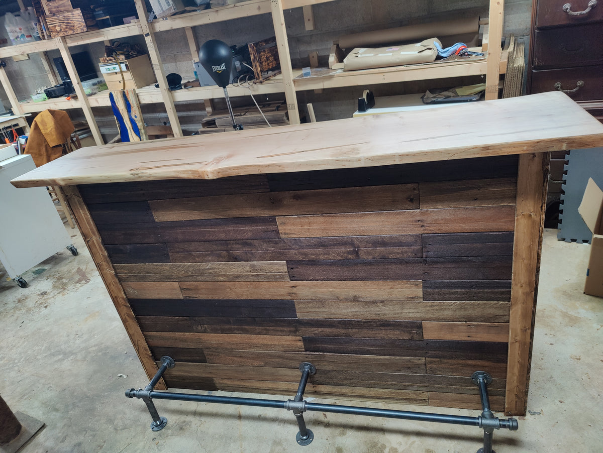 A rustic wooden bar counter with a live-edge top and a back panel made of mixed dark and medium brown wood planks, supported by a black industrial pipe base in a workshop setting.
