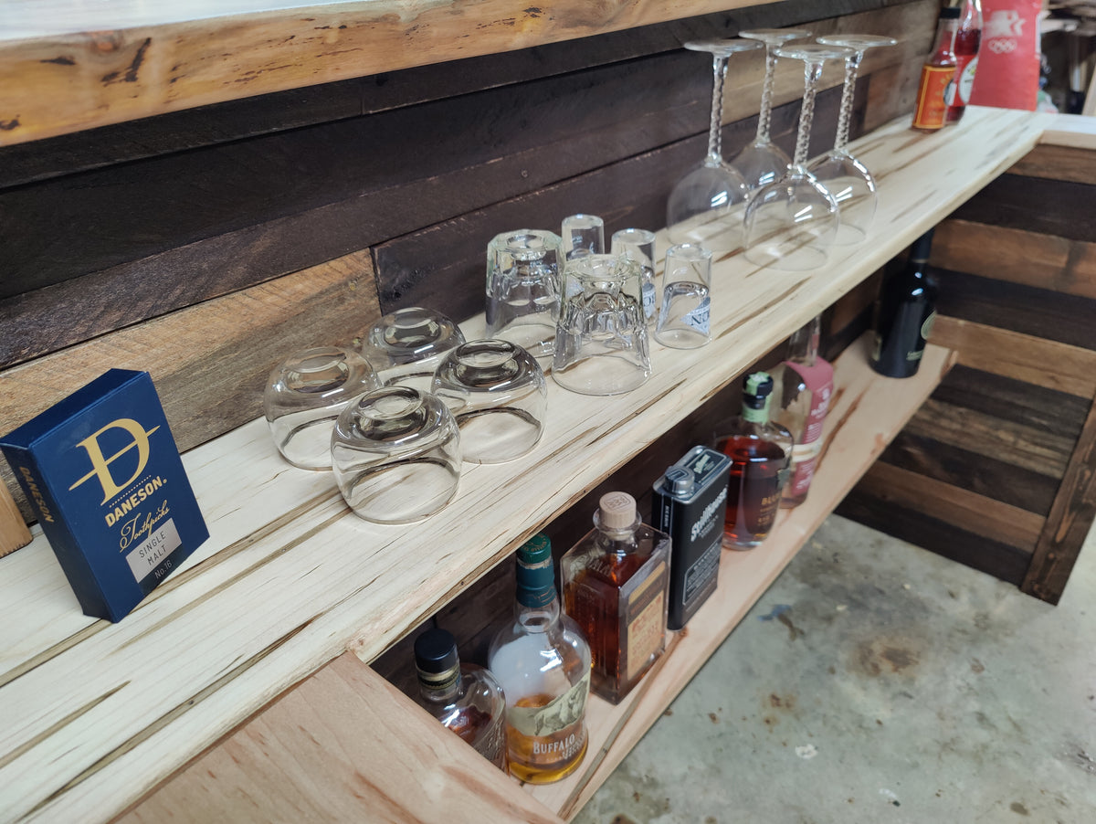 A rustic wooden bar shelf with multiple clear glass drinking glasses and several liquor bottles stored on the lower shelf. A blue box with a gold 'Damesh' logo sits on the left side of the shelf.