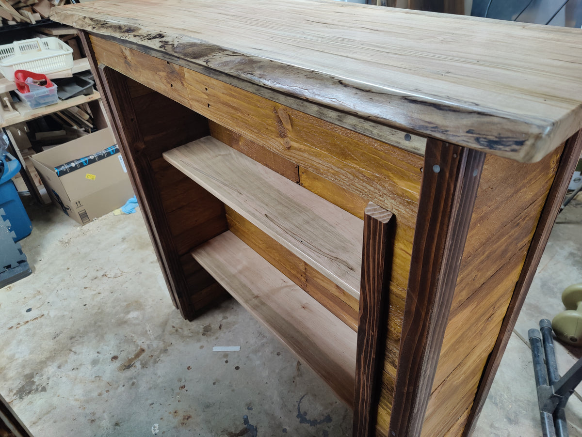 A rustic wooden shelving unit or cabinet with a live-edge natural wood top, featuring two open shelves and dark stained side panels, photographed in a workshop environment.