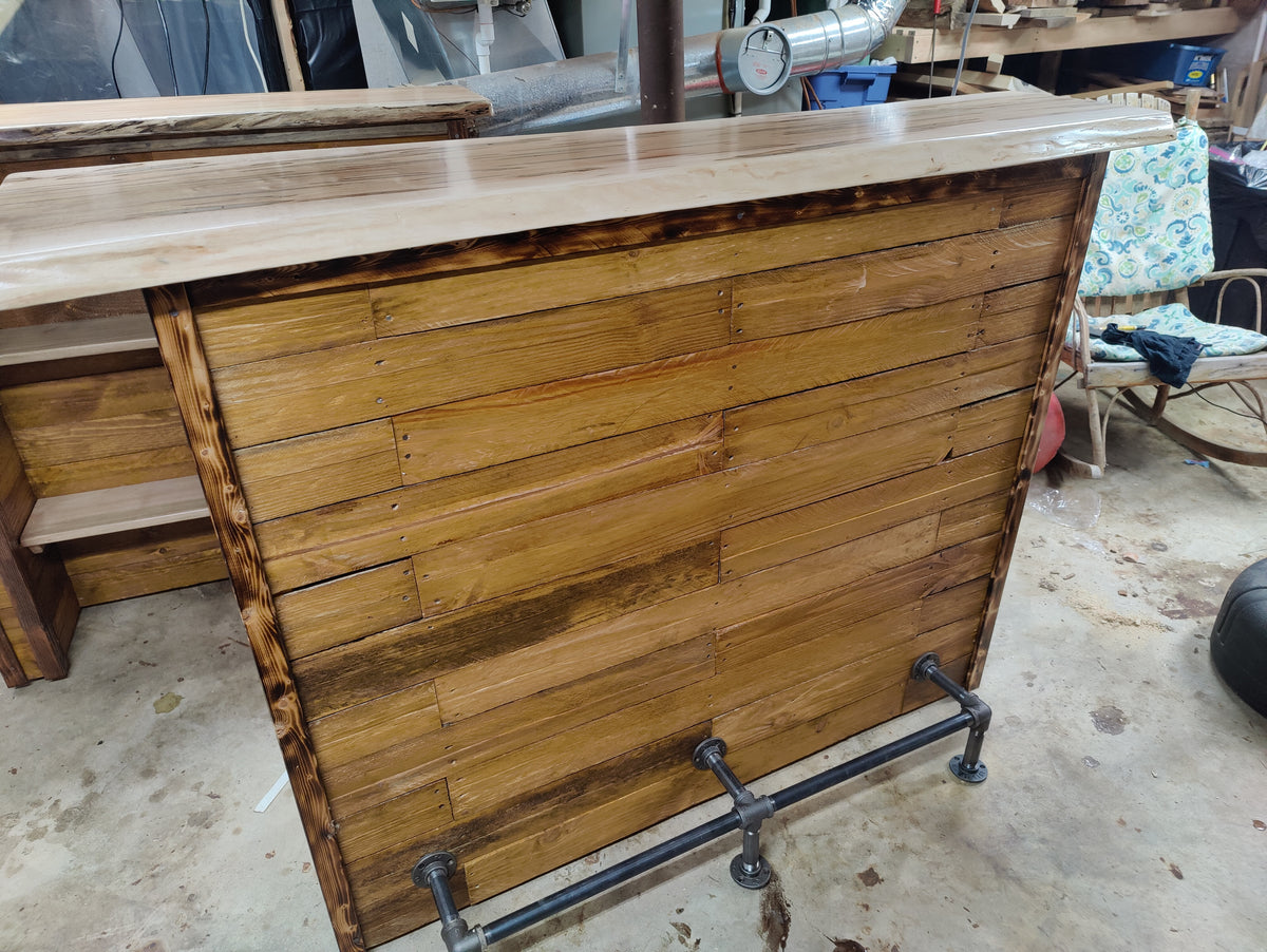 A rustic wooden bar counter with a slatted front and a dark metal pipe base, shown in a workshop setting.