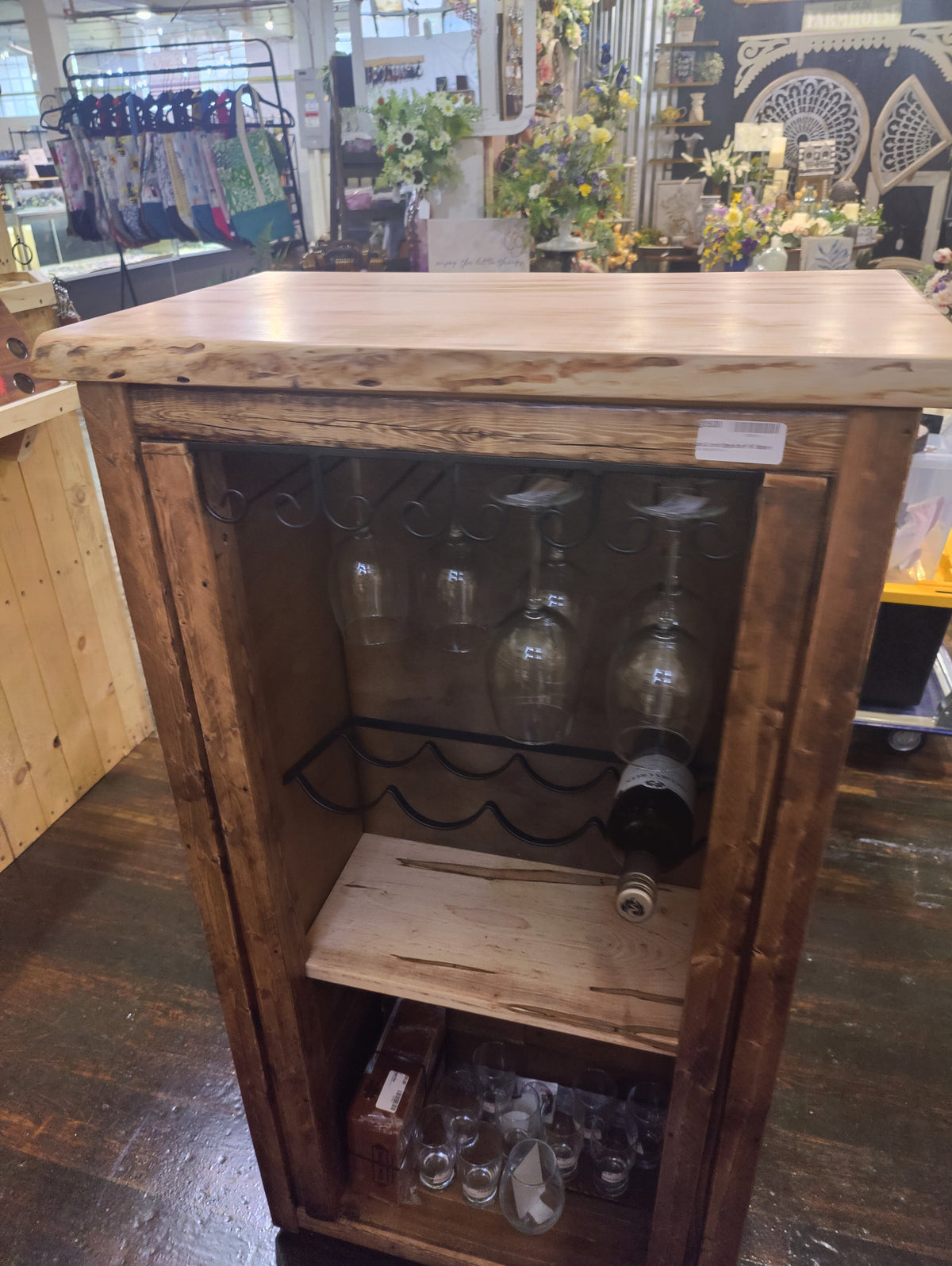 A rustic wooden wine cabinet with a thick natural wood top, a glass holder on the back panel, a wine rack, and two shelves, shown in a retail store setting.