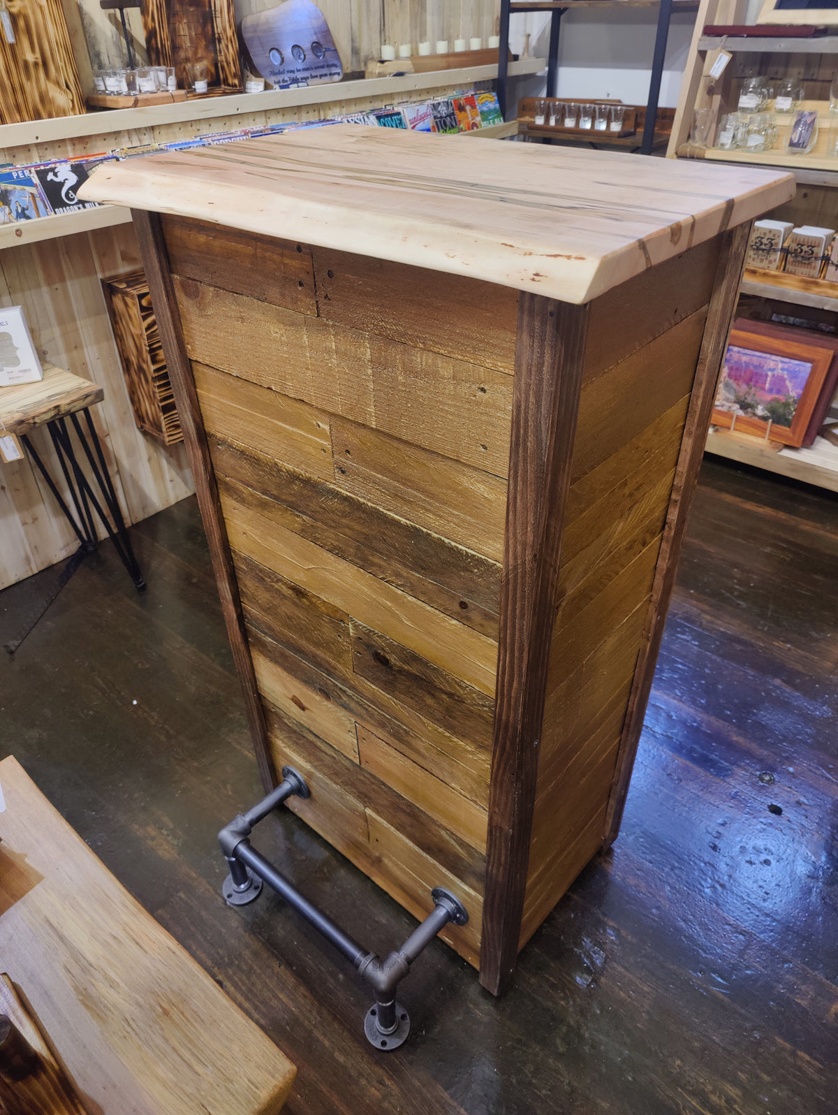 A rustic wooden bar counter or cabinet made from reclaimed wood planks, featuring a light natural wood top and dark stained side panels, with industrial pipe legs on a dark wood floor inside a shop.