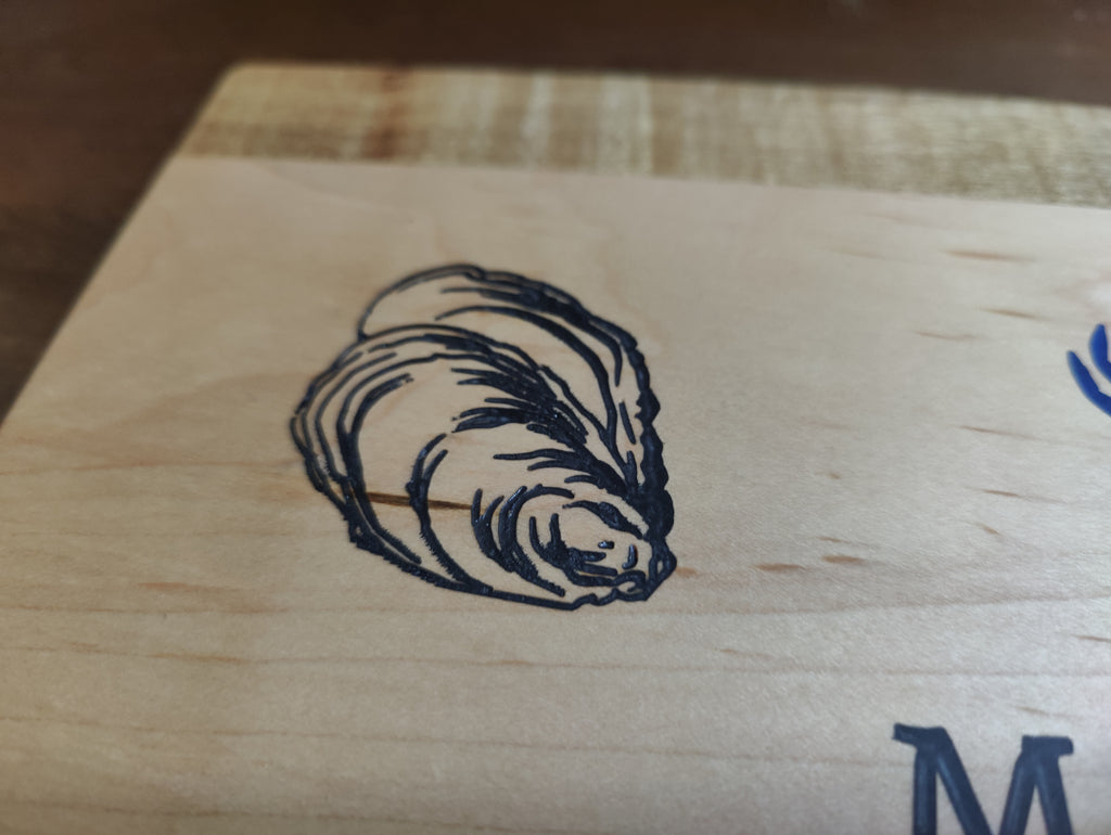 Close-up of a light natural wood surface with a dark engraved or printed illustration of a seashell and part of a blue letter or logo visible.