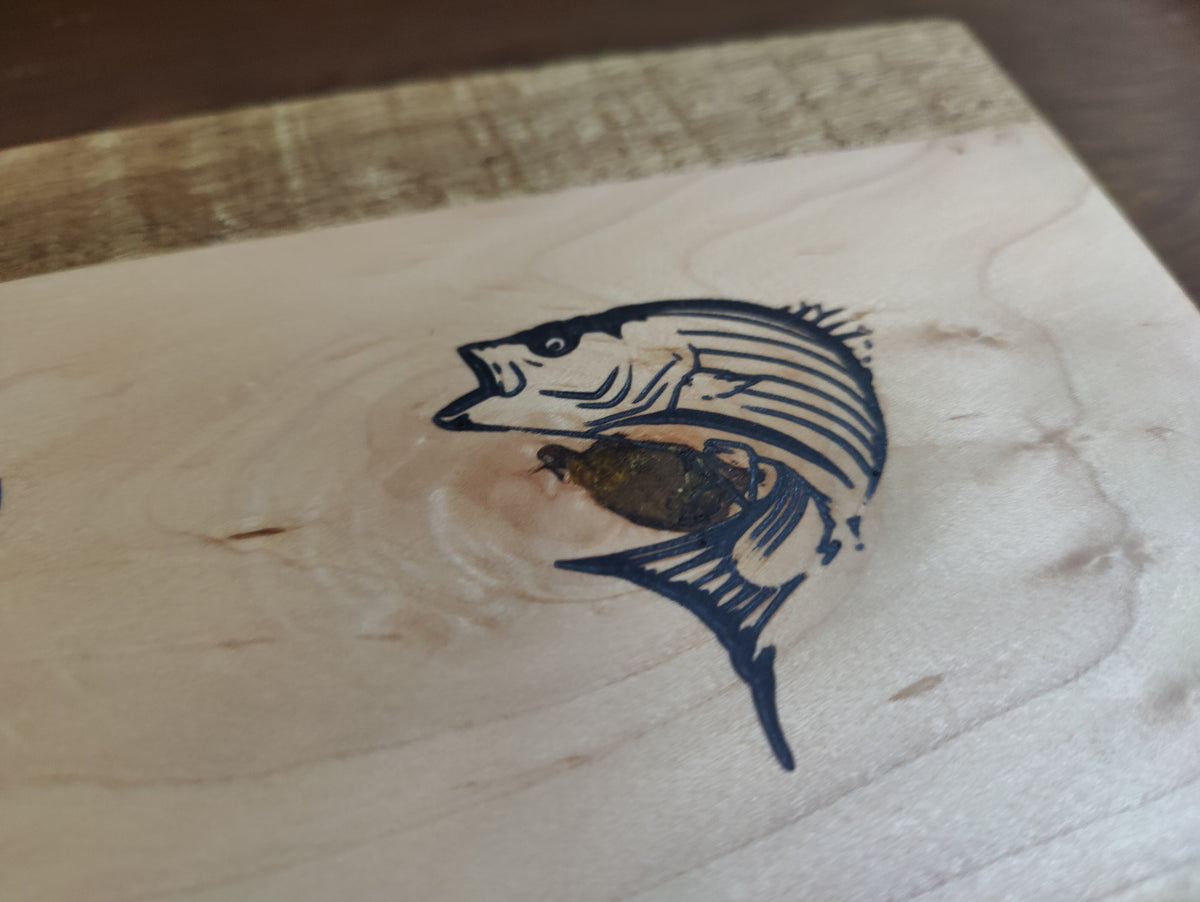A close-up of a light natural wood surface with a dark engraved or burned fish illustration in the center.
