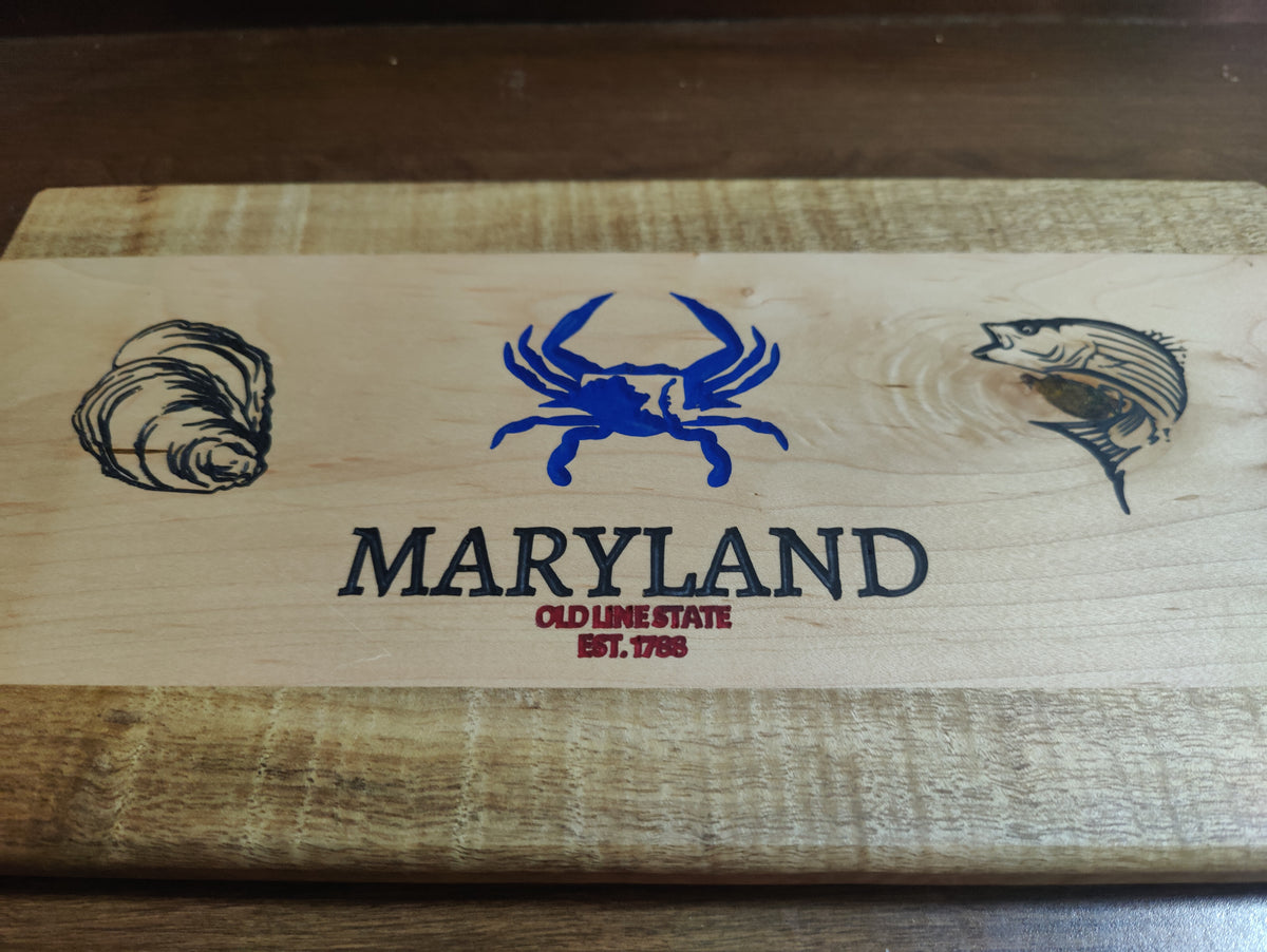 A rectangular wooden board or plaque with a light natural wood top and darker stained edges, featuring blue and black engraved graphics and text. The center shows a blue crab above the word 'MARYLAND' in black, with smaller red text reading 'OLD LIME STATE EST.