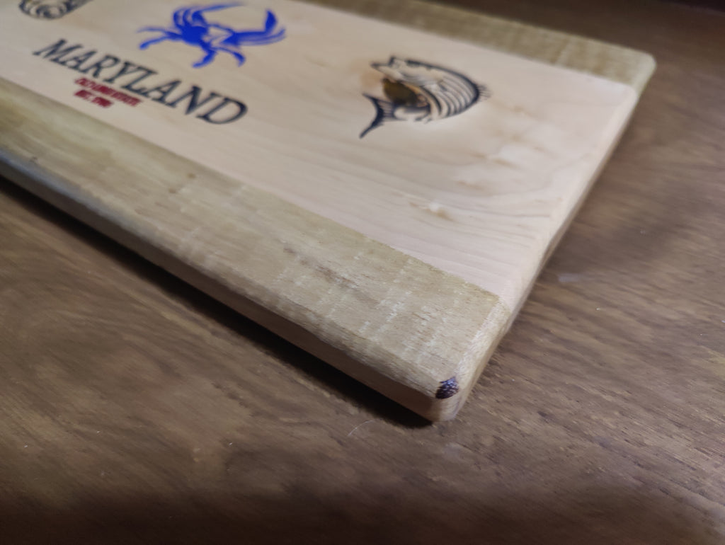 A close-up of a light natural wood cutting board resting on a wooden table. The board features a blue Maryland crabs logo and a black engraved fish design.