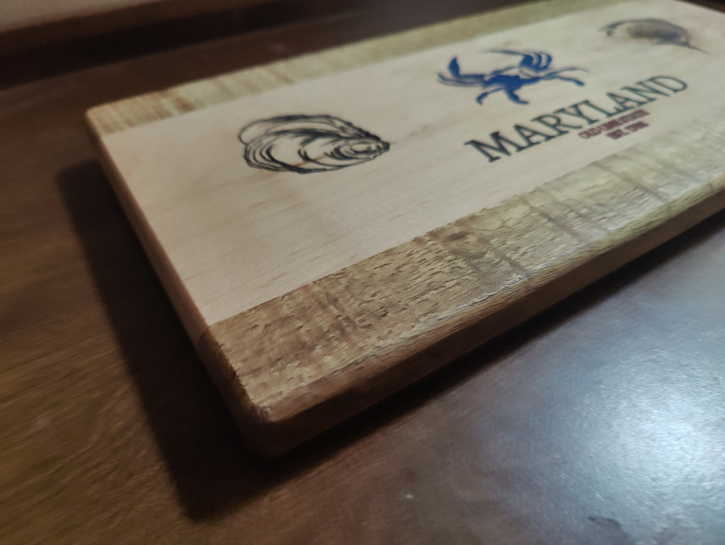 A rectangular wooden cutting board with a natural wood grain finish, featuring a blue crab logo and the word 'MARYLAND' engraved on the top, resting on a dark wooden table.