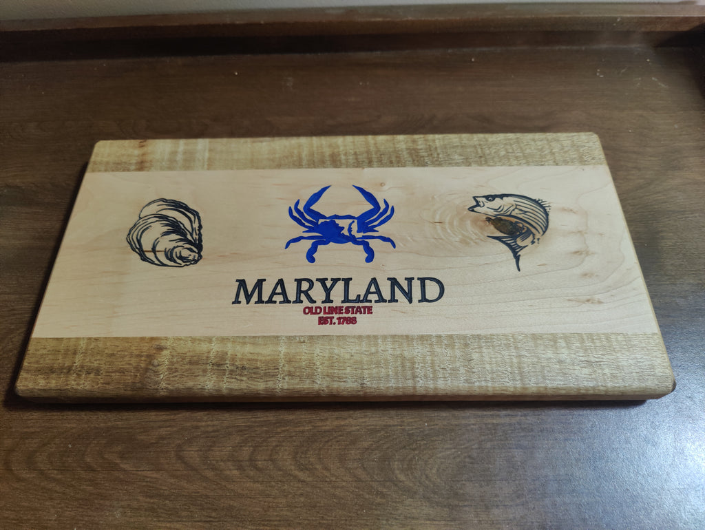 A rectangular wooden cutting board with a natural wood grain finish, featuring a blue crab, oyster shell, and fish engraving, with 'MARYLAND' and 'OCEAN FISH EST. 2016' printed in the center.