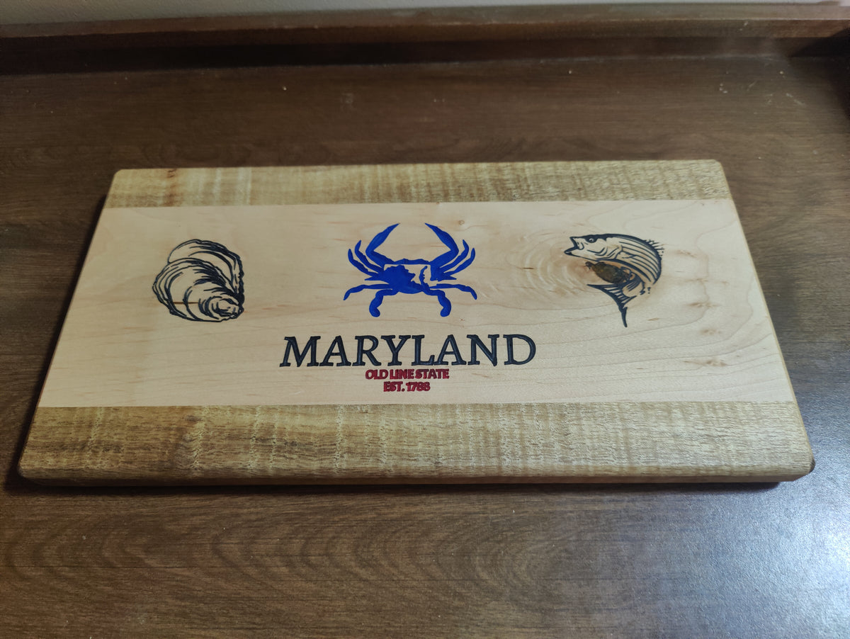 A rectangular wooden cutting board with a natural wood grain finish, featuring a blue crab, oyster shell, and fish engraving, with 'MARYLAND' and 'OCEAN FISH EST. 2016' printed in the center.