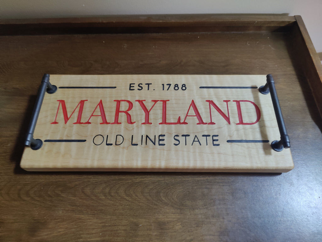 A rectangular wooden sign with black metal pipe-style handles, featuring the words 'EST. 1788 MARYLAND OLD LINE STATE' in red and black lettering, displayed on a dark wood surface.