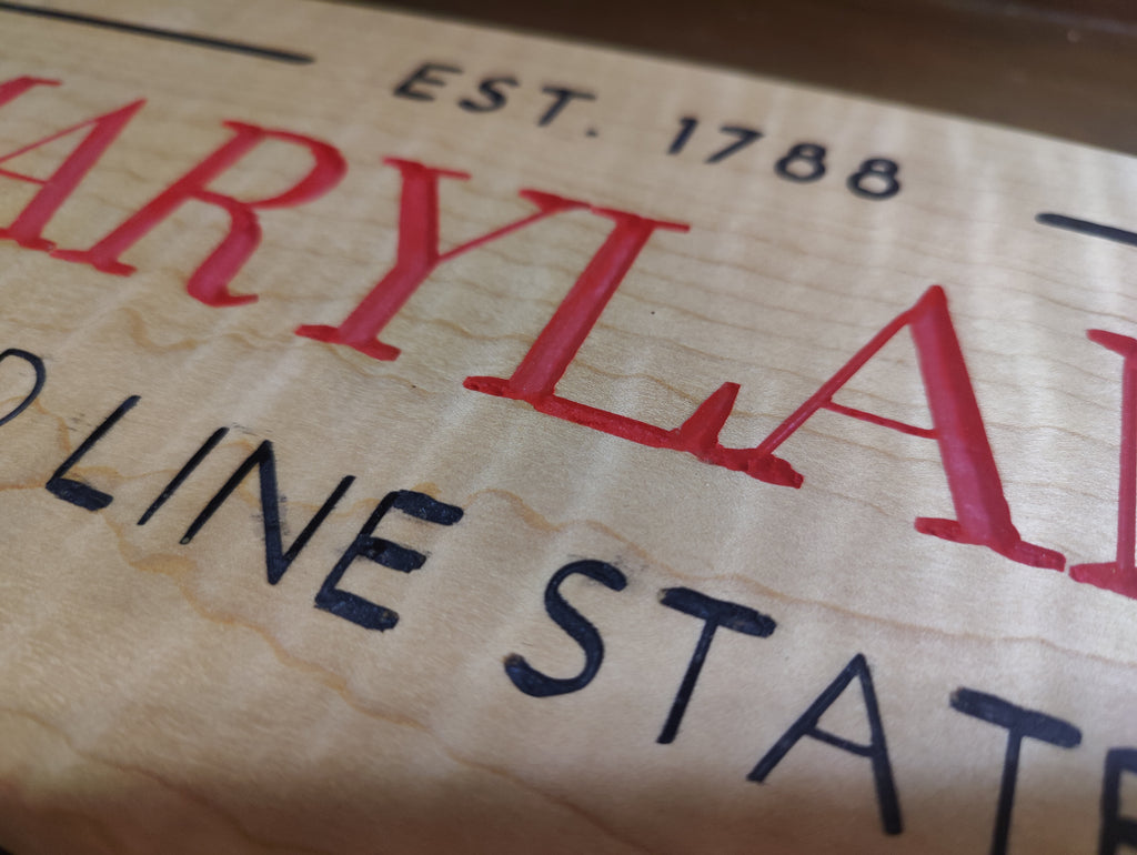 Close-up of a wooden sign featuring red and black text on a light wood grain background, including 'MARYLAND', 'EST. 1788', and 'LINE STATE'.