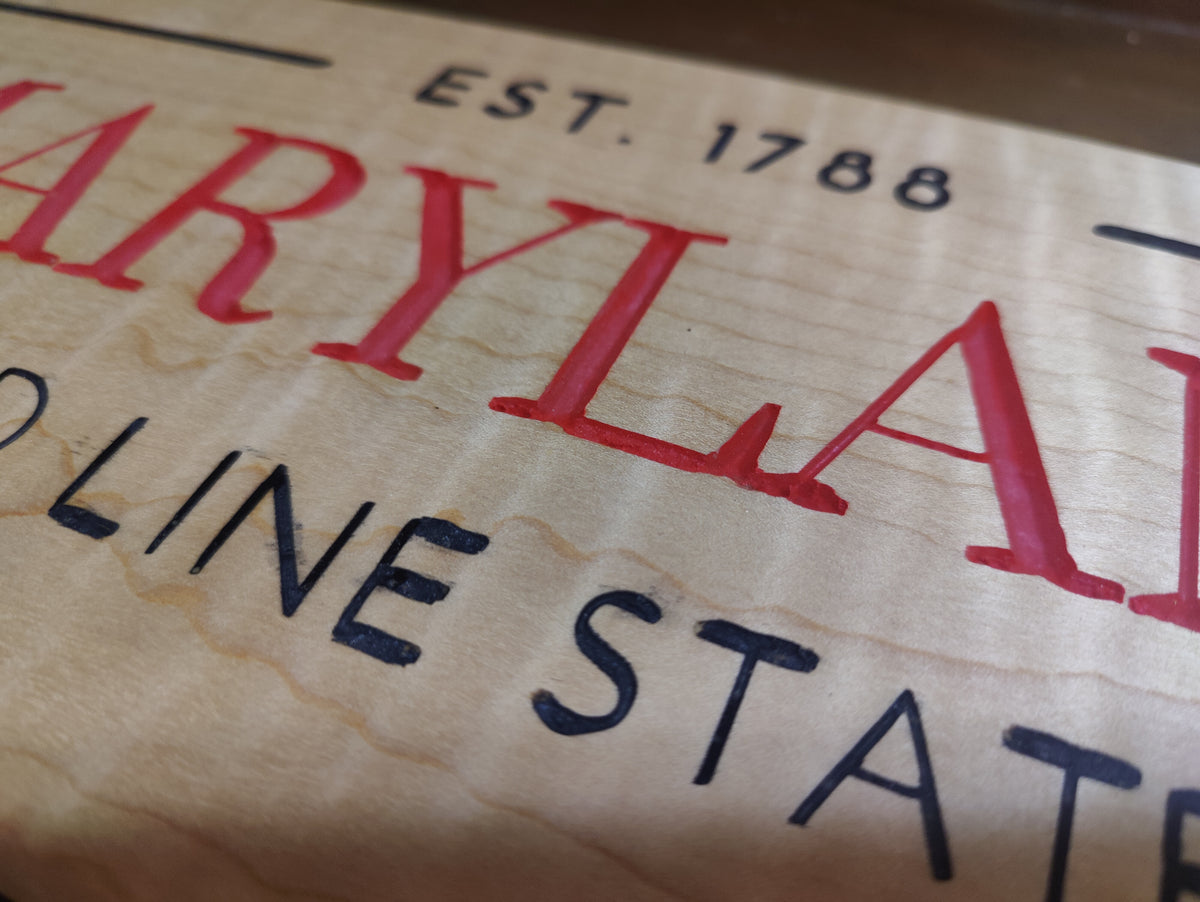 Close-up of a wooden sign featuring red and black text on a light wood grain background, including 'MARYLAND', 'EST. 1788', and 'LINE STATE'.