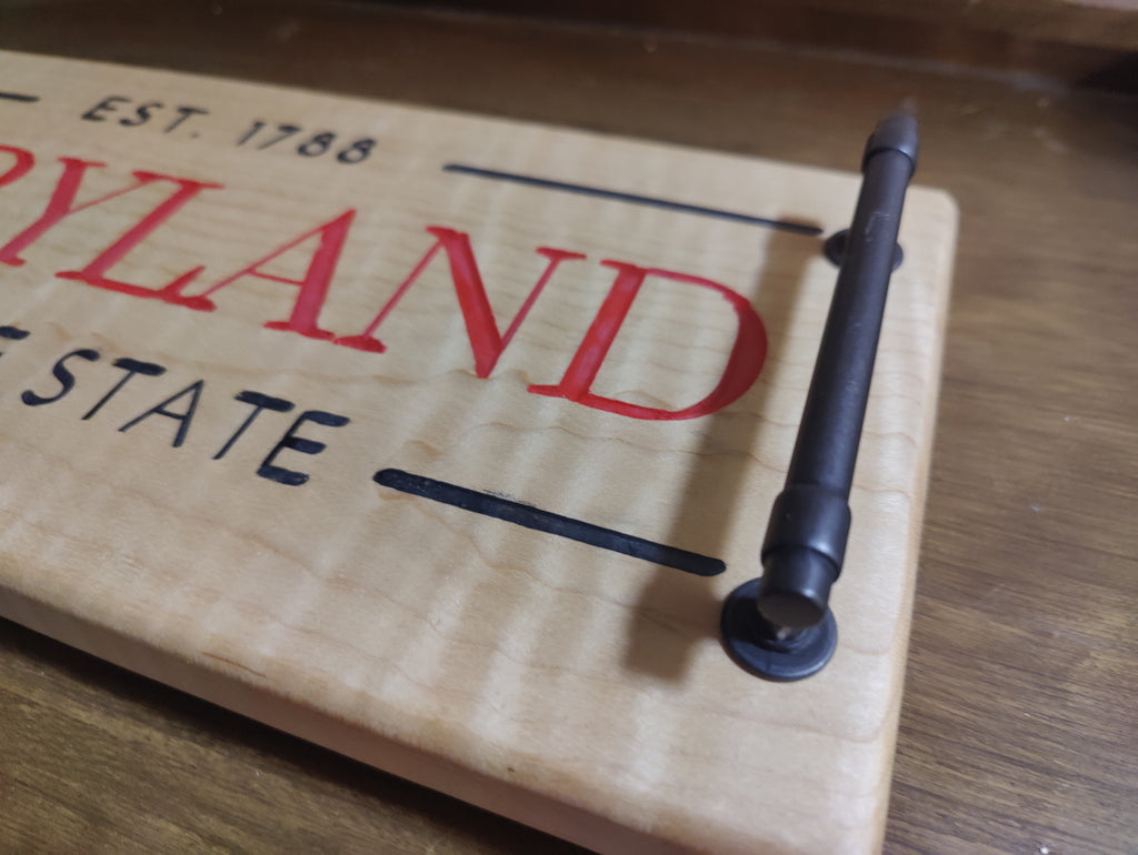 A close-up of a wooden sign featuring Maryland state text in red and black, with a dark metal nail mounted on the wood.