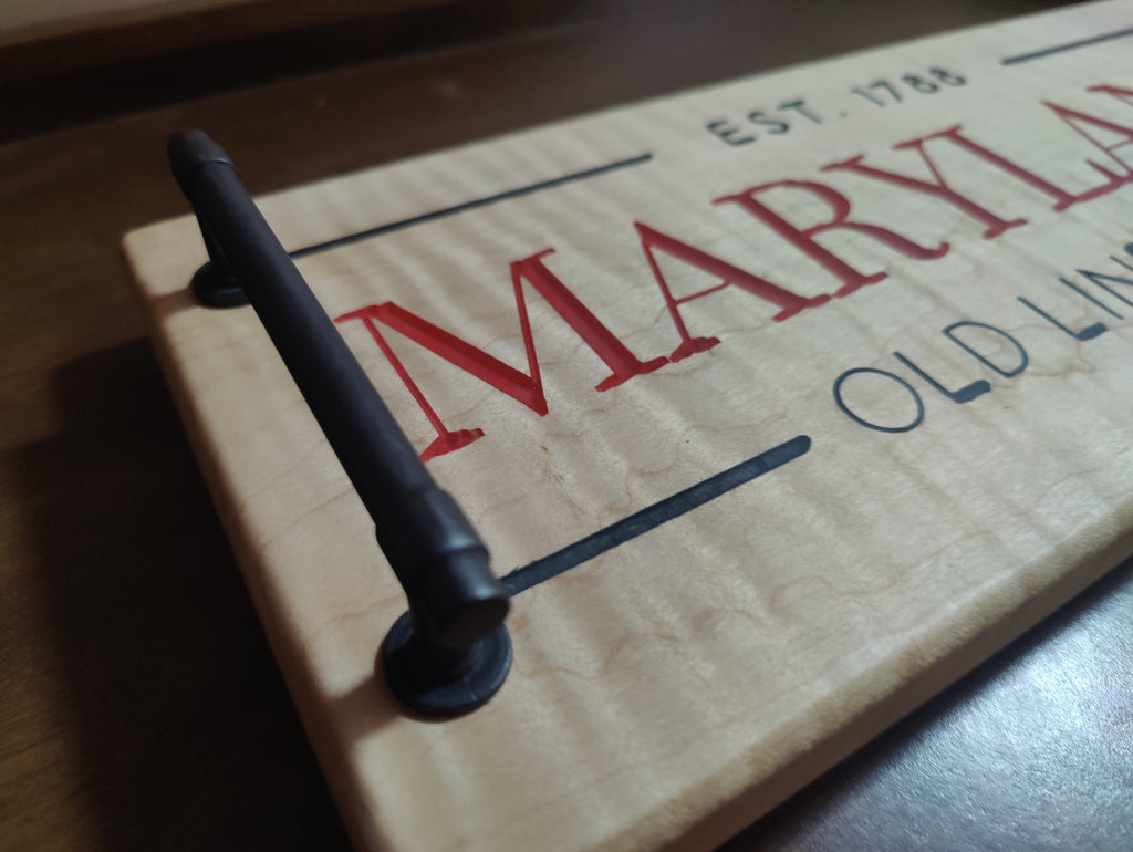 Close-up of a rectangular wooden sign with a black metal handle, featuring red and blue text that reads 'MARYLAND' and 'EST. 1788' on a dark wooden surface.