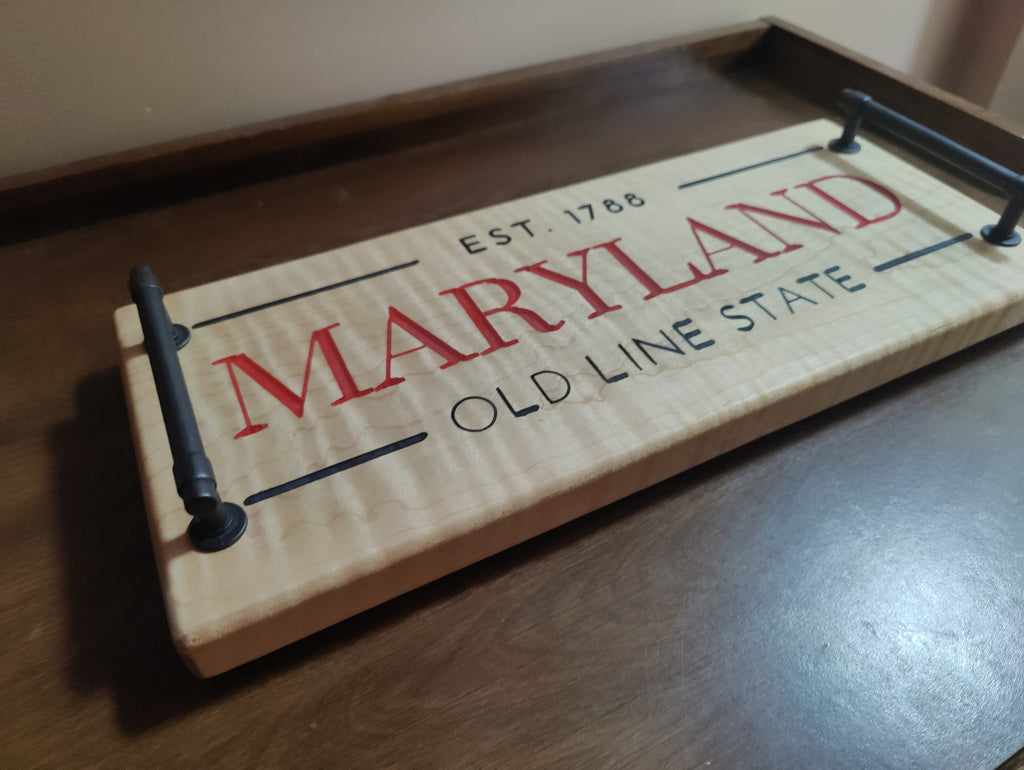A rectangular wooden sign with black metal handles, featuring 'MARYLAND OLD LINE STATE EST. 1786' printed on a light wood surface, resting on a dark wooden table.