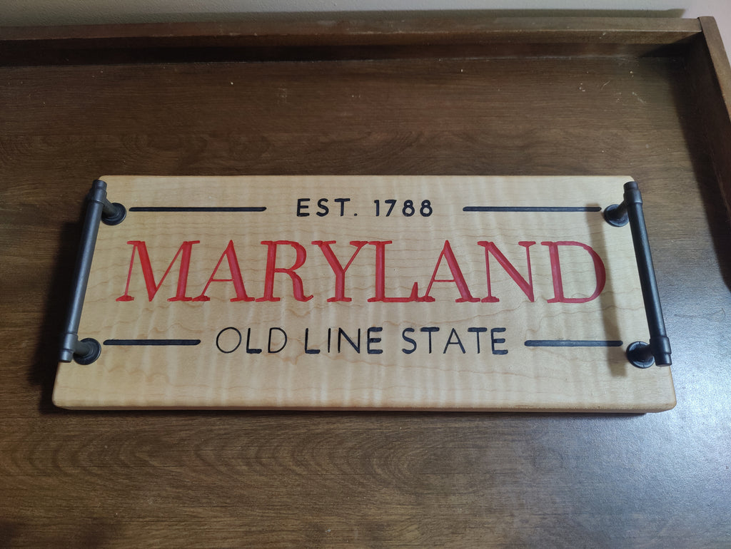 A rectangular wooden sign with black metal corner brackets and handles, featuring 'EST. 1788 MARYLAND OLD LINE STATE' printed on a light wood background, resting on a dark wooden surface.