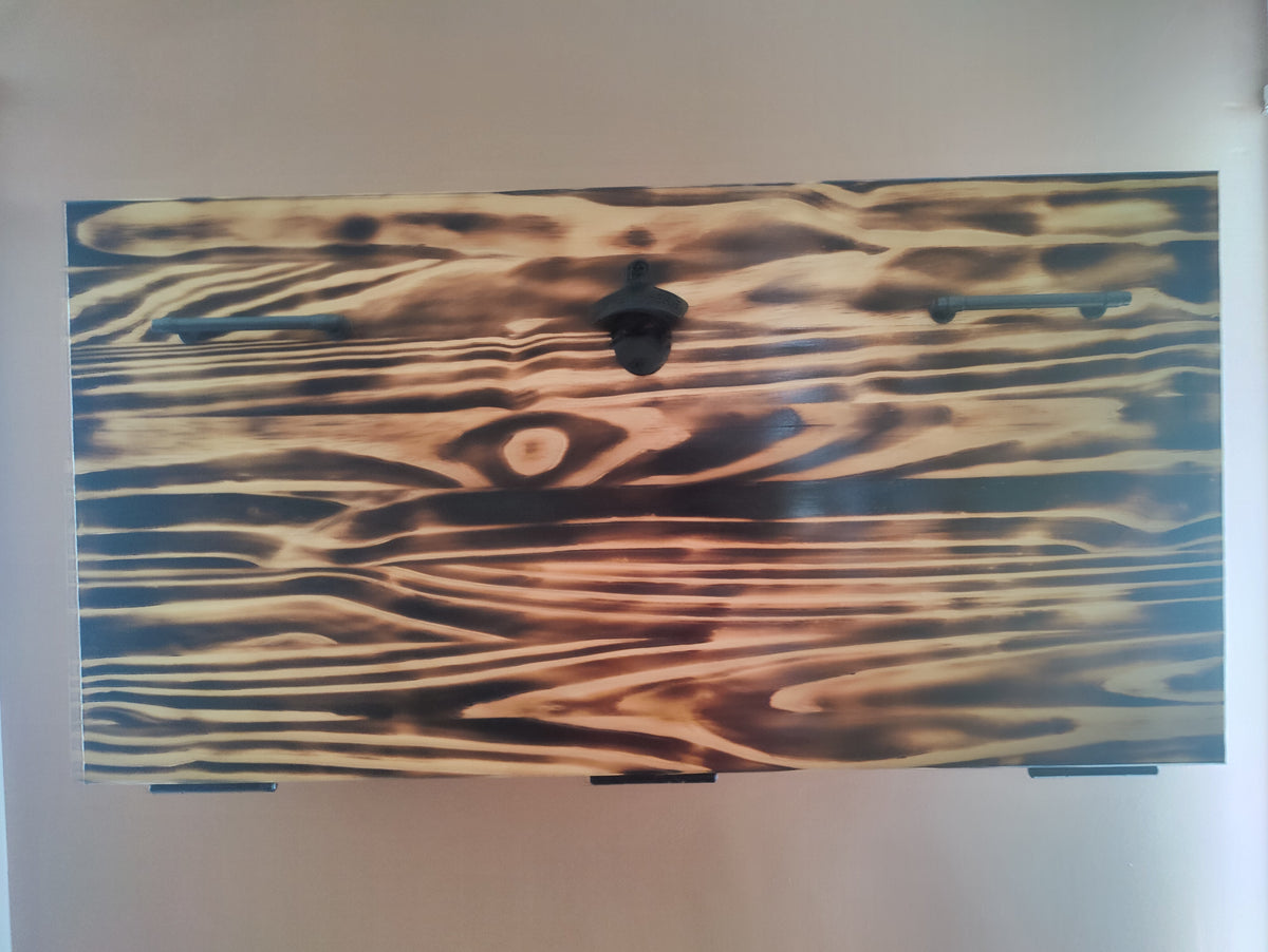 A rectangular wall-mounted artwork featuring a stylized wood grain or flame-like pattern in black, brown, and tan tones, shown against a plain light wall.