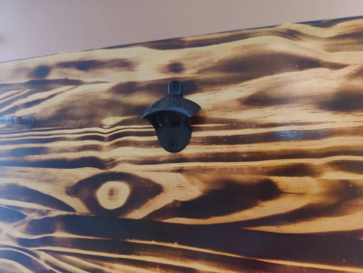 A close-up of a rustic wooden surface with a dark metal bottle opener mounted on it. The wood has a charred, burnt finish showing warm brown and black tones with visible grain patterns.