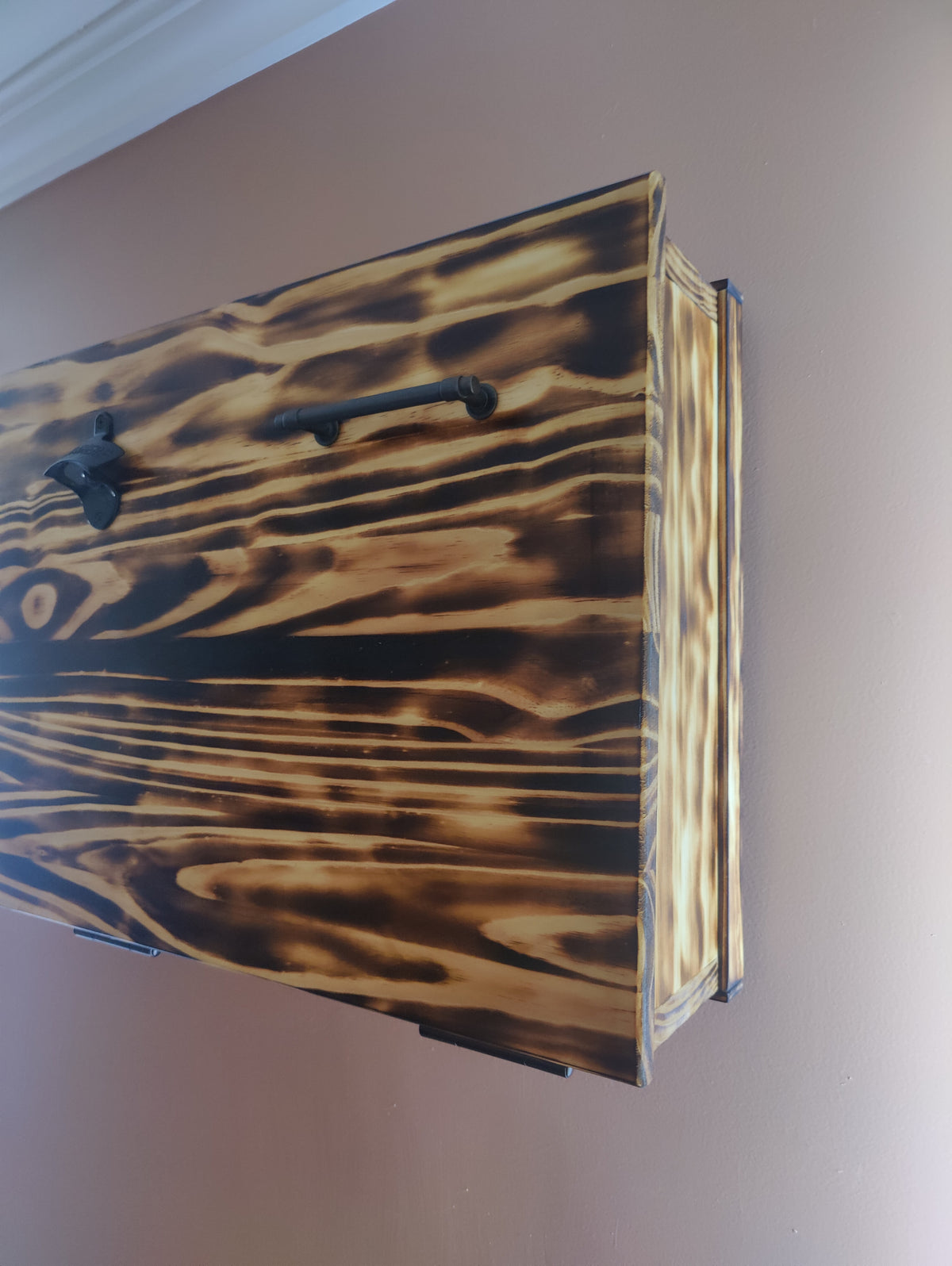 A rectangular wooden cabinet or wall-mounted box with a dark brown and black charred wood grain finish, featuring a black metal handle and mounted against a beige wall.
