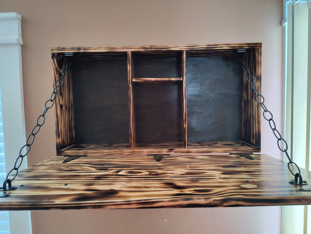 A rustic, handcrafted wooden wall-mounted shelf or desk hanger suspended by black metal chains. The wood features a dark brown finish with prominent black charred or burnt wood grain patterns, set against a plain beige wall.