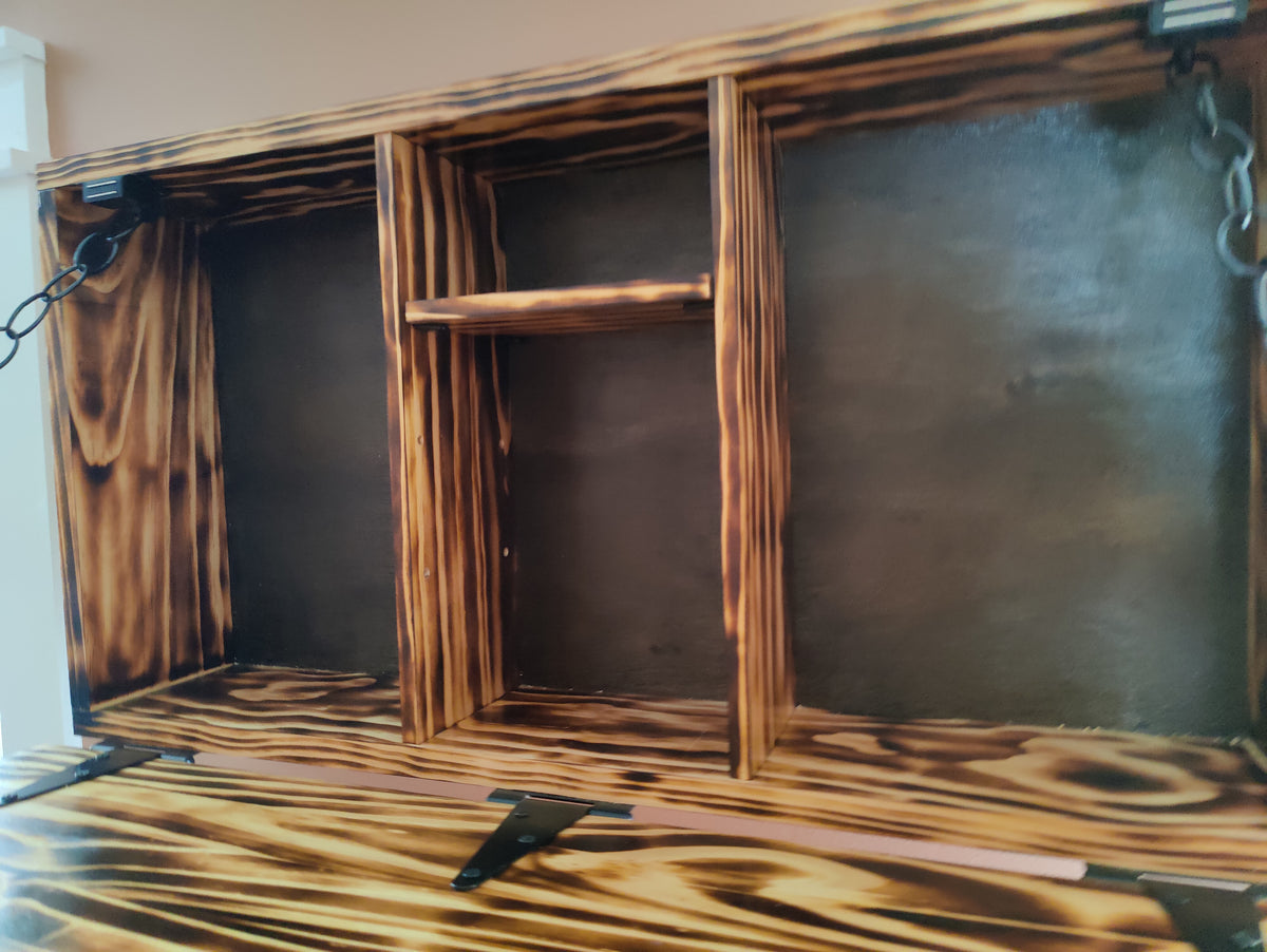 A rustic, handcrafted wooden wall-mounted cabinet or shelf with three open compartments, featuring a dark brown and black charred wood finish, metal hanging brackets, and a hinged front panel on a wooden surface.