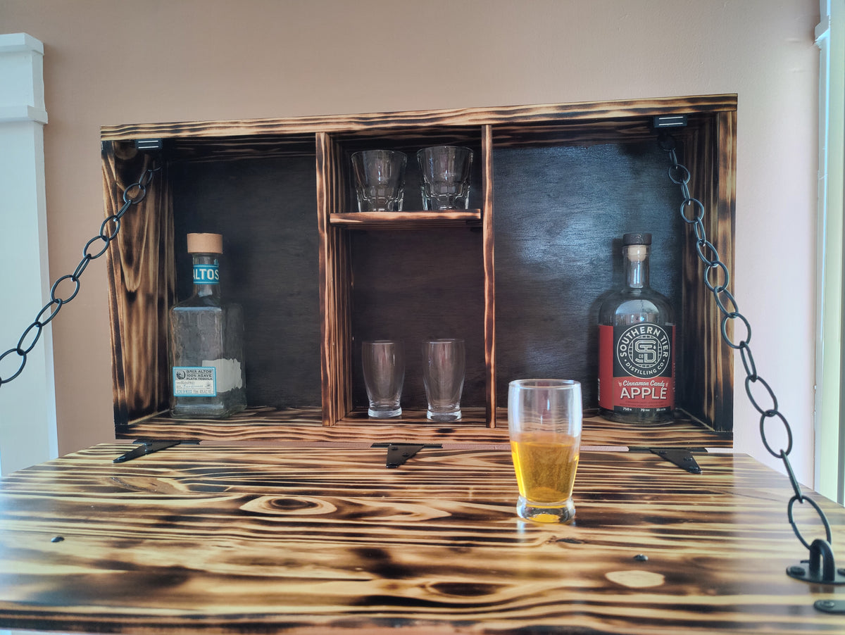 A rustic wooden bar hutch with a dark stained finish, featuring three open shelves, hanging chains, and a matching wooden tabletop. A glass of beer and bottles are placed on and inside the hutch.