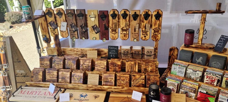 A rustic wooden display stand filled with engraved wooden signs, small wooden boxes, and packaged goods at what appears to be an outdoor market or booth. The display includes various engraved wood pieces with animal and landscape designs, along with boxed items and packaged products.