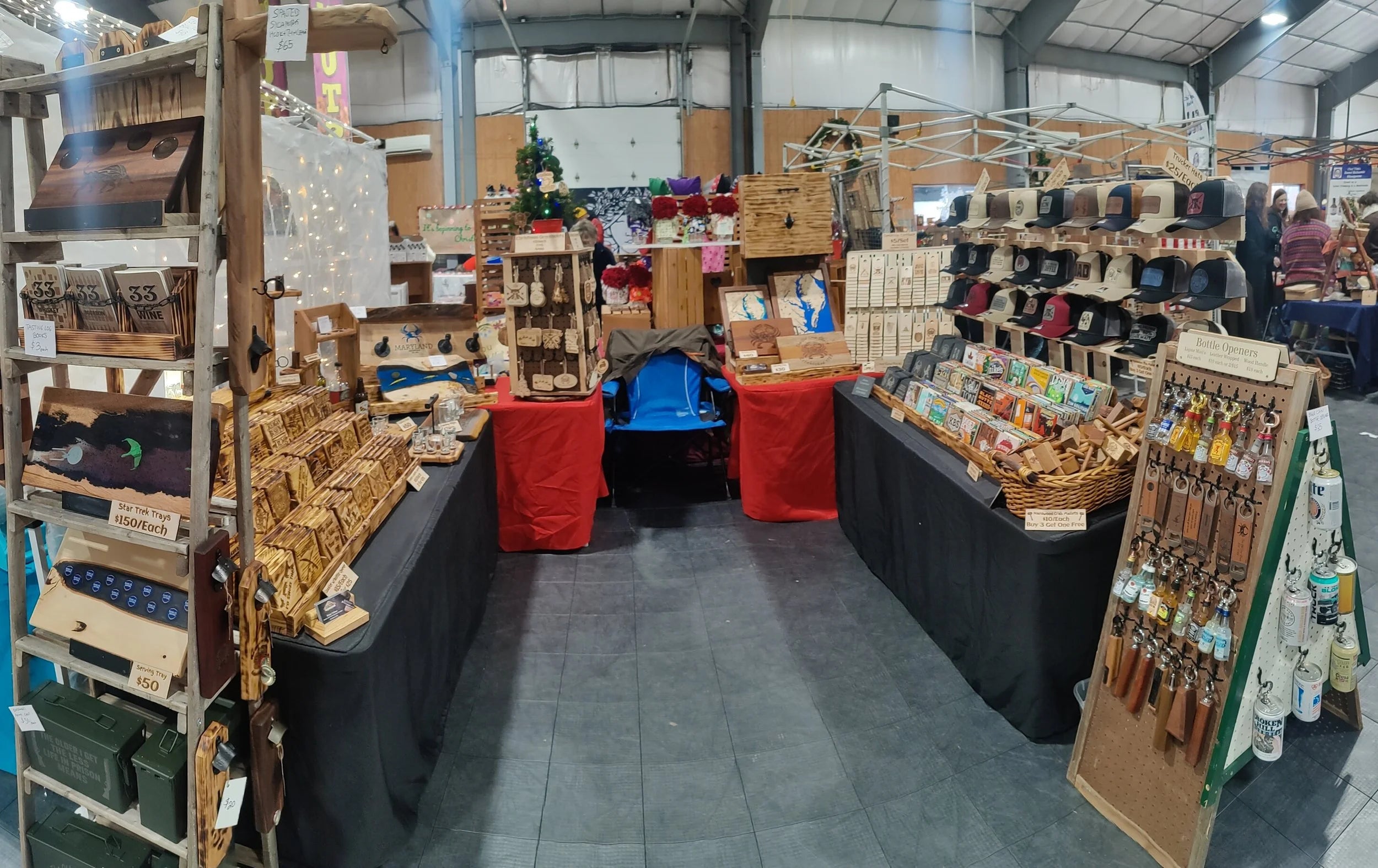 A wide view of a market stall inside a warehouse or fairground, displaying various handmade wooden crafts, baskets, tools, and packaged goods on black-clothed tables and wooden shelving.