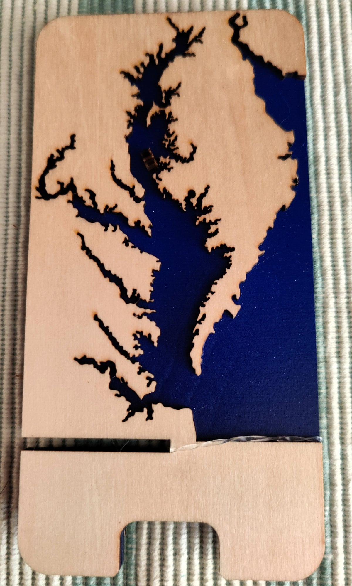 A wooden phone stand with a laser-cut map design of a river delta, featuring a natural light wood finish and a dark blue painted section, resting on a textured gray woven fabric.