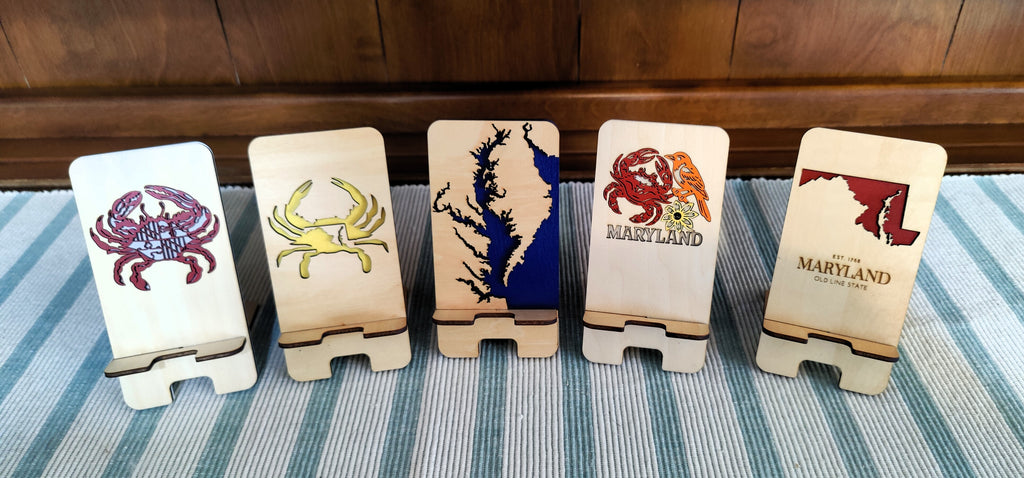 Five wooden coast marker stands arranged in a row on a blue and white striped blanket, with a dark wood headboard in the background. Each stand features a different Maryland-themed design, including crabs, a map silhouette, and the Maryland state outline with text.