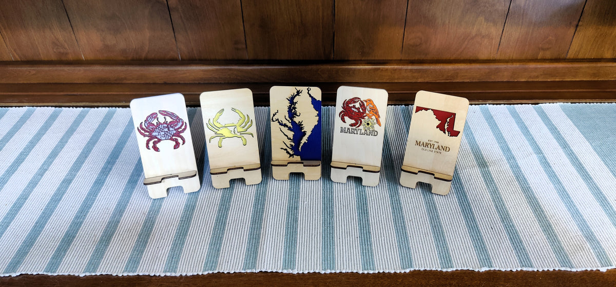 Five wooden Maryland state-themed coasters arranged in a row on a blue and white striped runner, with a dark wood panel background.