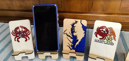 Three wooden phone stands with Maryland-themed designs are displayed on a striped blue and white surface in front of a wooden wall. One stand features a red crab, one features a blue and black map design, and one features a red crab with 'MARYLAND' text.