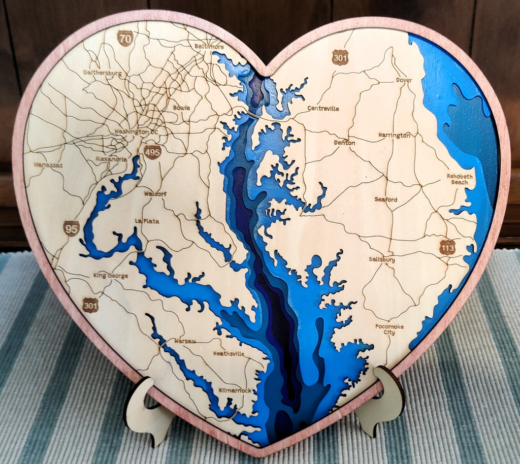 Heart-shaped wooden map plaque of the Chesapeake Bay, featuring laser-cut geography with blue water layers and a light natural wood background, displayed on a striped surface.