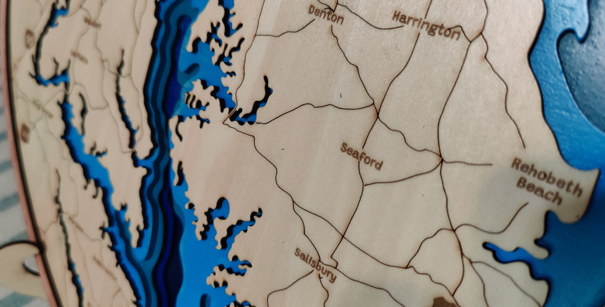 Close-up of a wooden map showing coastal geography with cutout blue water areas and engraved place names including Seaford, Rehoboth Beach, and Karrington.
