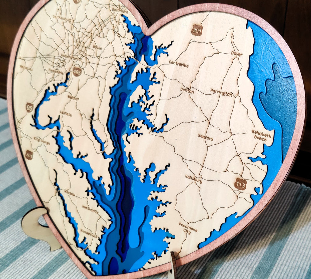 Heart-shaped wooden map plaque of the Chesapeake Bay, featuring layered blue water levels and engraved city names on a light wood background, displayed on a striped surface.