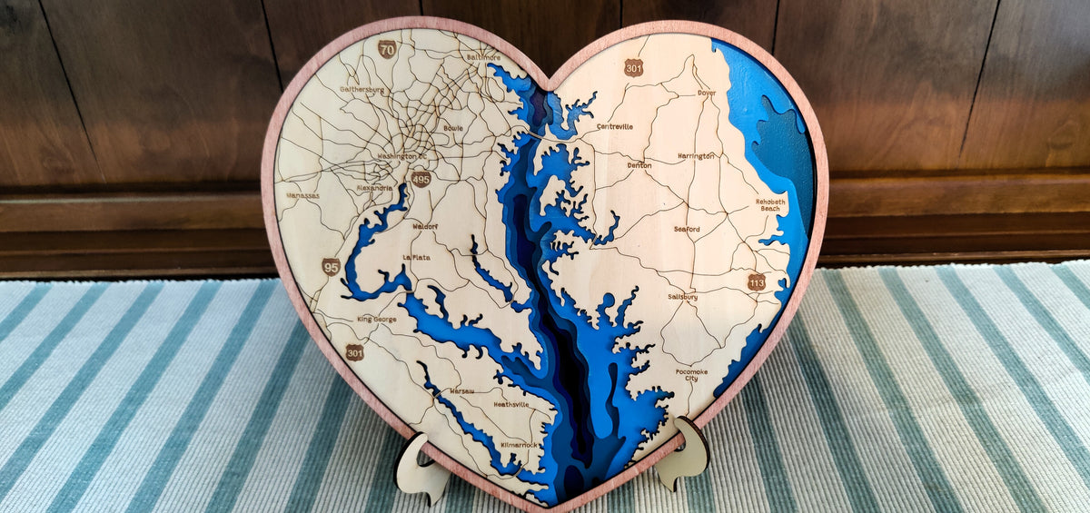 Heart-shaped wooden map plaque of the Chesapeake Bay, featuring detailed etched coastline and waterways with blue resin fill, displayed on a striped rug in front of a dark wood panel background.