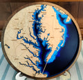 A round wooden plaque featuring a stylized map of the Chesapeake Bay with layered blue water effects and engraved place names, displayed on a small stand.