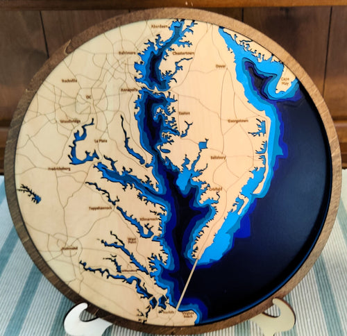 A round wooden plaque featuring a stylized map of the Chesapeake Bay with layered blue water effects and engraved place names, displayed on a small stand.