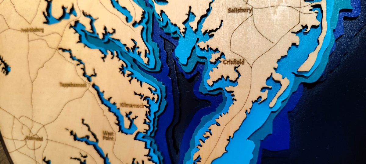 Close-up of a wooden layered depth map plaque showing coastal waterways and place names such as Salisbury and Crisfield, with wood grain and cutouts in beige, tan, brown, and blue tones.