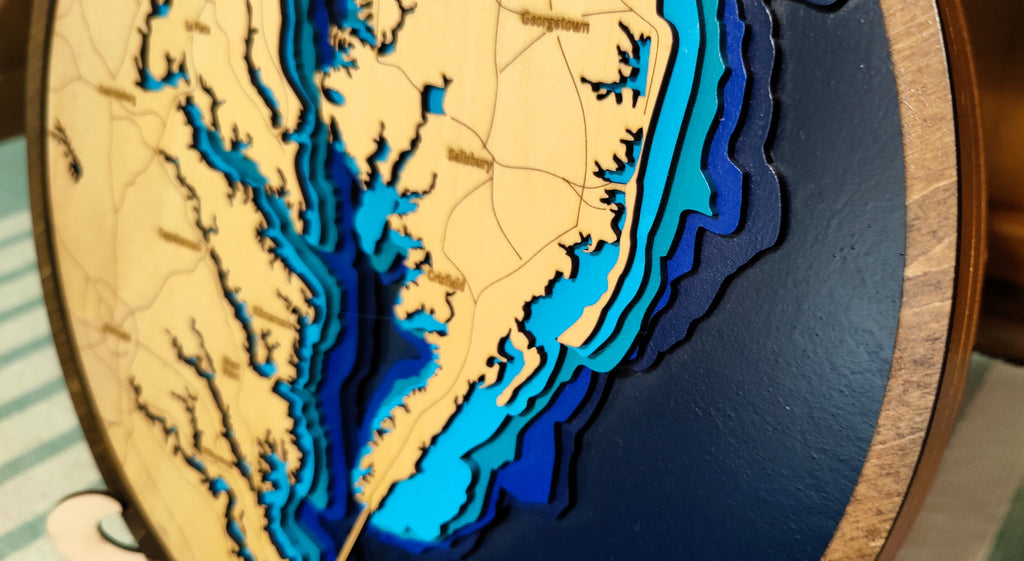 A close-up of a circular wooden 3D laser-cut map plaque showing the coastline of North Carolina and surrounding areas, with layered wood sections in beige, light blue, medium blue, and dark blue.