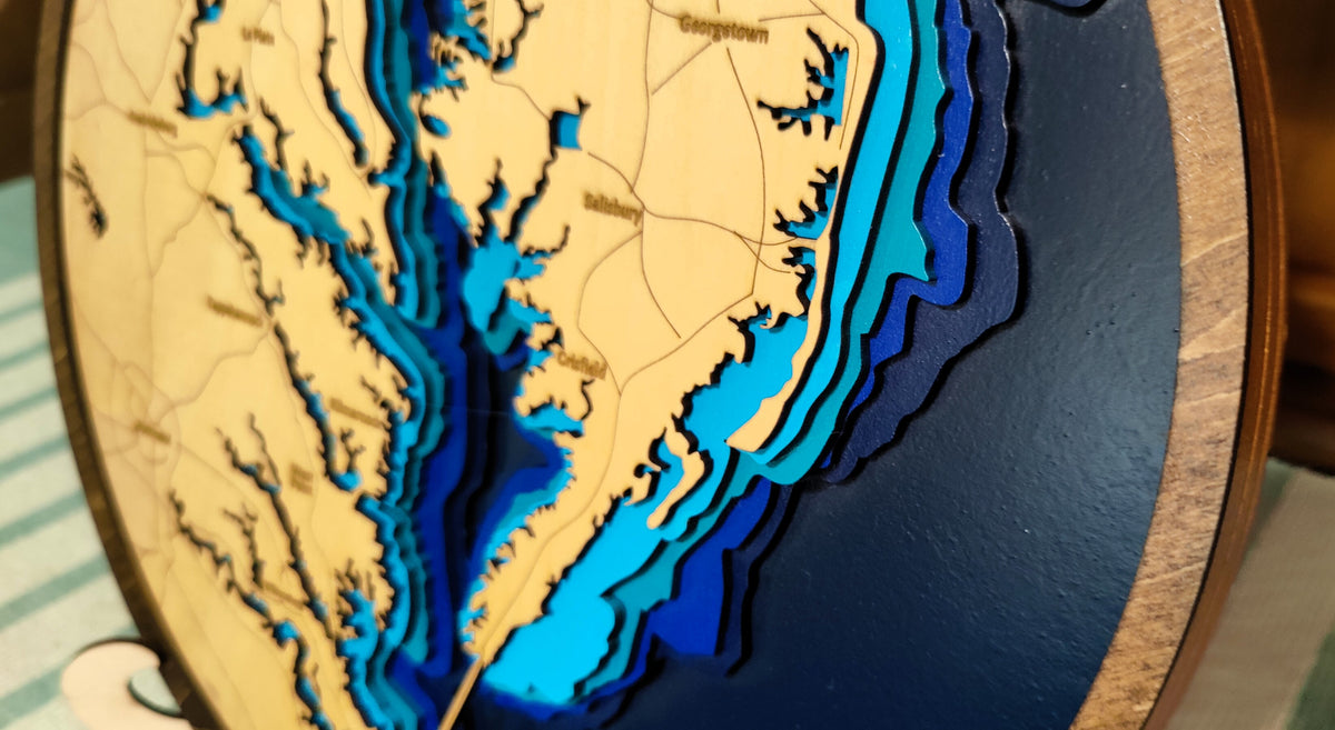 A close-up of a circular wooden 3D laser-cut map plaque showing the coastline of North Carolina and surrounding areas, with layered wood sections in beige, light blue, medium blue, and dark blue.