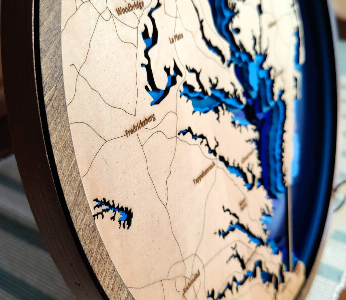Close-up of a circular wooden map plaque showing a lake and river system with labeled places such as Woodbridge, Upton, and Freehold, in layered beige wood with dark blue water shapes and a dark wood rim.