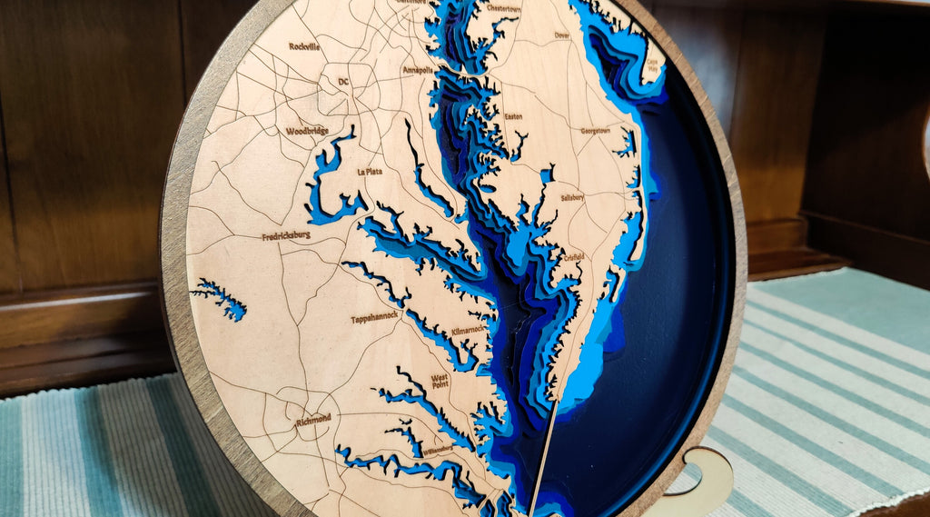 A circular wooden depth-map wall art piece showing the Chesapeake Bay watershed with layered blue and navy water effects and engraved place names, photographed indoors against a dark wood background.