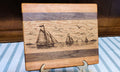 A rectangular wooden cutting board with a laser-engraved nautical scene of sailboats on the water, displayed on a small metal stand over a blue and white striped tablecloth.