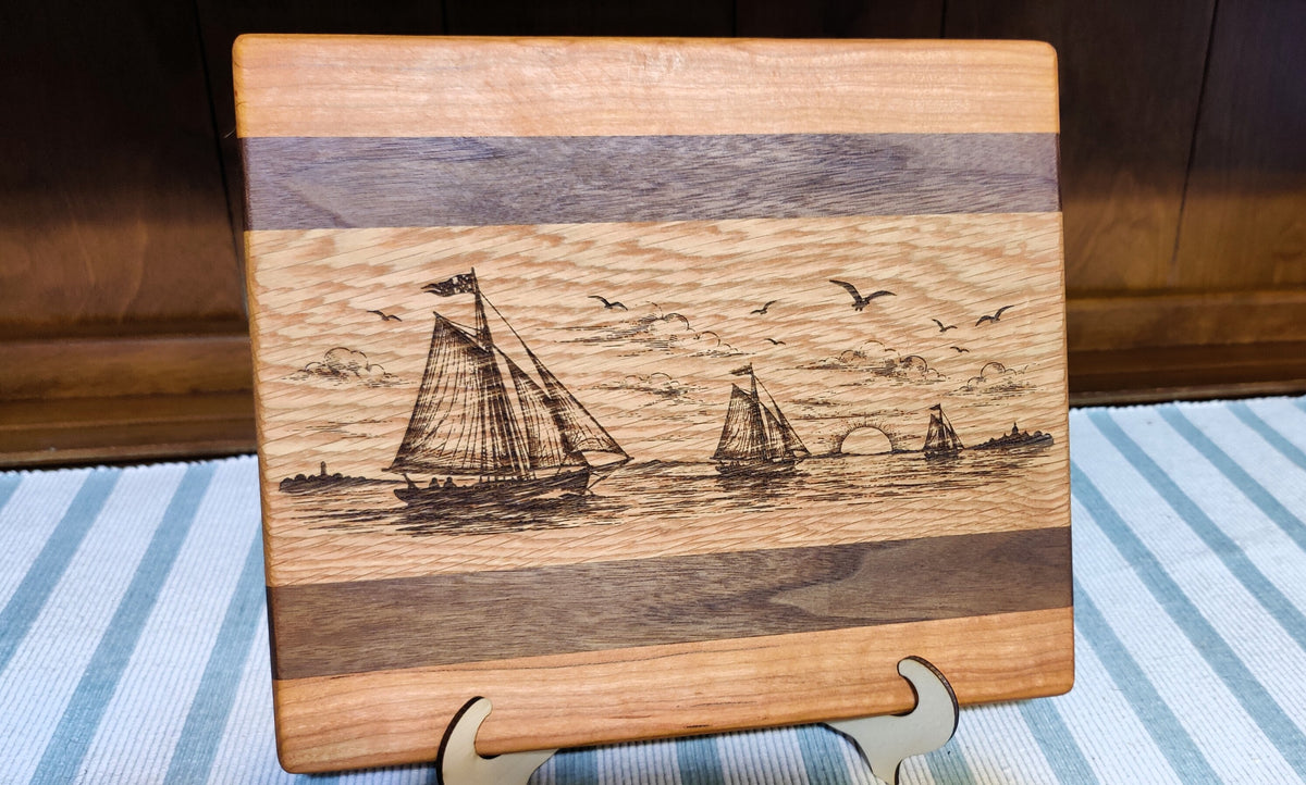 A rectangular wooden cutting board with a laser-engraved nautical scene of sailboats on the water, displayed on a small metal stand over a blue and white striped tablecloth.